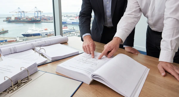 Plan serré professionnel montrant deux consultants en tenue professionnelle examinant ensemble un dossier technique complexe ouvert sur une table, avec des documents contractuels, cahiers des charges et classeurs administratifs dispersés alentour. En arrière-plan visible par la fenêtre, un port maritime avec navires et installations portuaires. Lumière naturelle professionnelle, atmosphère de collaboration et d'expertise technique dans un contexte maritime authentique. Seuls les mains, documents et environnement de port visible doivent être en focus.